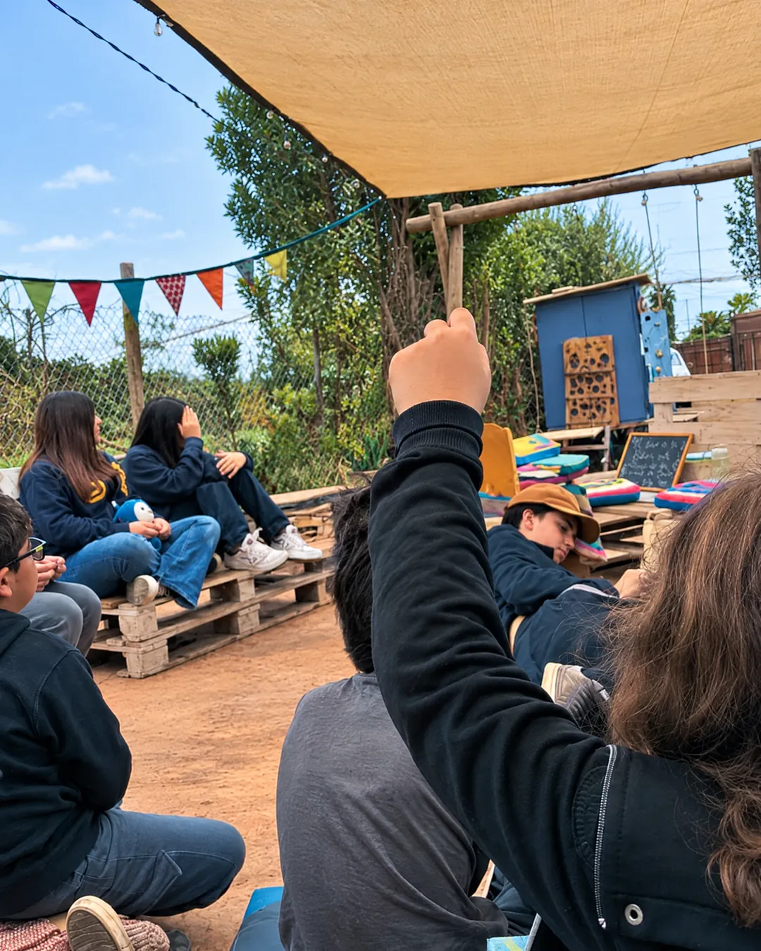 Jóvenes conversando sentados en bancas de pallets bajo un toldo
